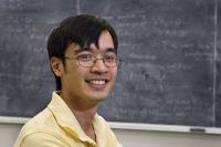 Zdjęcie uśmiechniętego Terence'a Tao na tle zapisanej tablicy. 